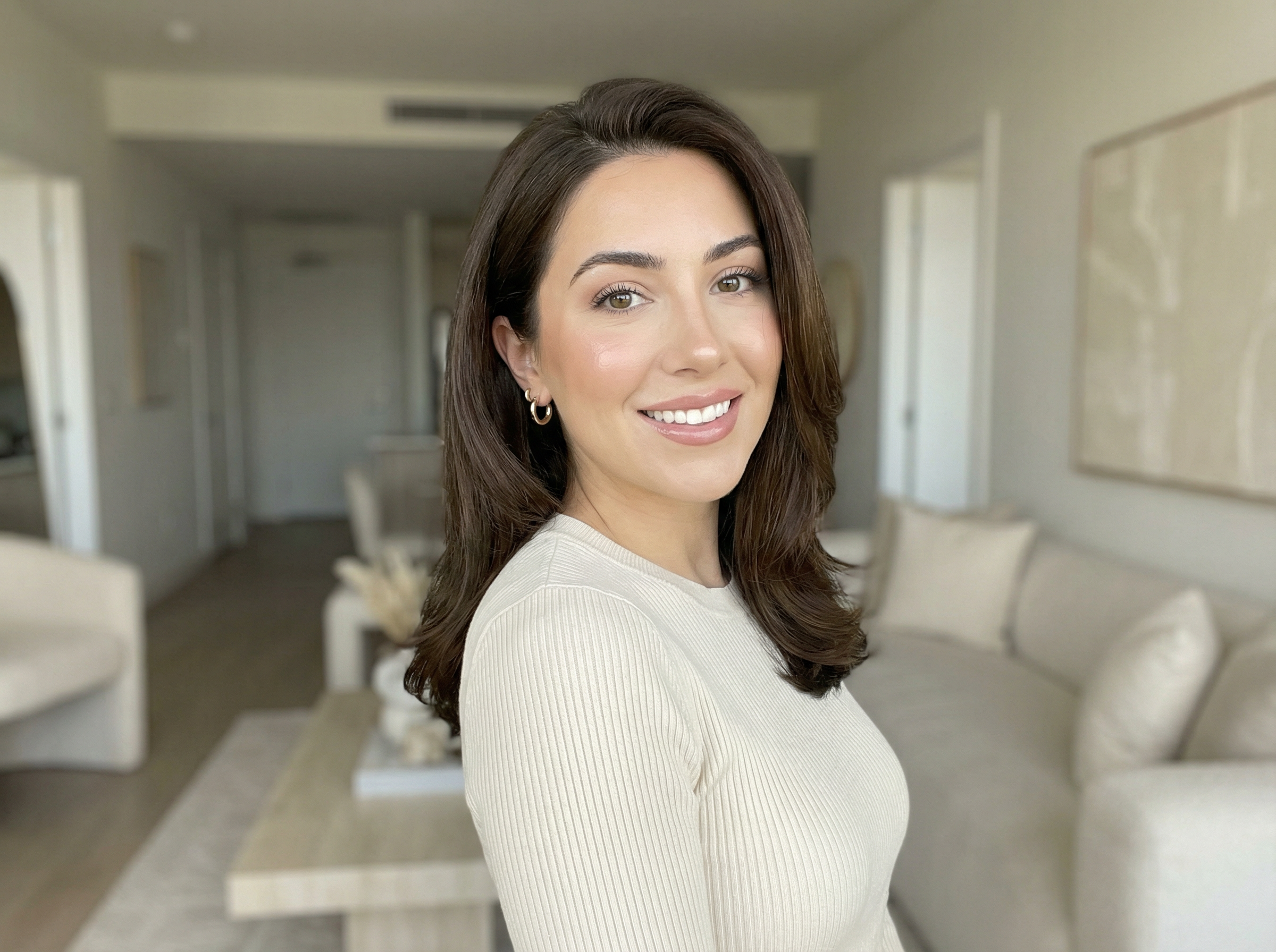 Profile-ready portrait of a fictional beauty influencer with glossy shoulder-length waves, soft glam makeup, and a cream knit top in a bright neutral apartment interior.